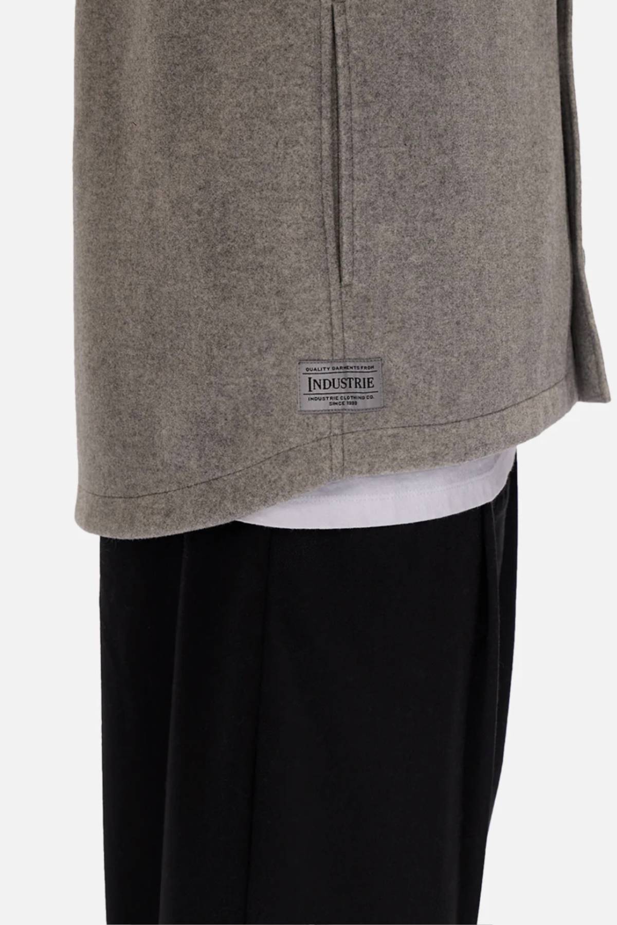 Close-up of a person wearing the relaxed fit Industrie The New Coleman Jacket in light gray, with a classic collar and small "INDUSTRIE" label by the hem, layered over a white shirt and black pants. Only the lower torso and upper legs are shown.
