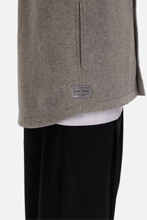 Close-up of a person wearing the relaxed fit Industrie The New Coleman Jacket in light gray, with a classic collar and small "INDUSTRIE" label by the hem, layered over a white shirt and black pants. Only the lower torso and upper legs are shown.