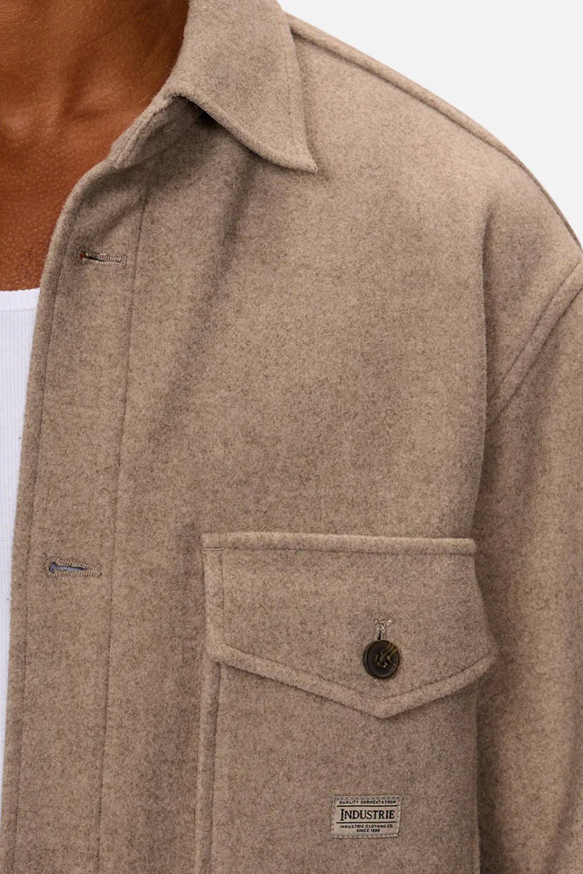 Close-up of a person wearing the Industrie The New Coleman Jacket in beige wool with a classic collar, twin chest pockets (one with an "Industrie" label), and visible metal button, styled over a white ribbed shirt.