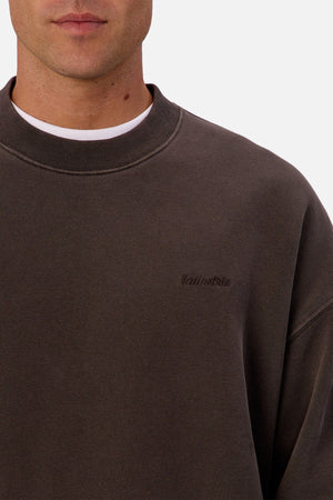 A close-up of a person wearing the dark brown Industrie The New Del Sur Sweatshirt, layered over a white t-shirt. The embroidered logo is visible on the left chest, highlighting its premium construction. Only their chin and lower face are shown.
