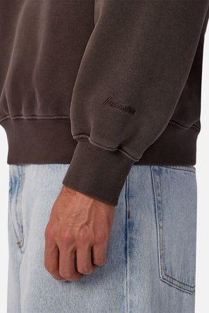 A person wears the Industrie The New Del Sur Sweatshirt in oversized dark brown with subtle branding near the wrist, paired with light blue jeans and shown from the waist down with their left hand relaxed at their side.