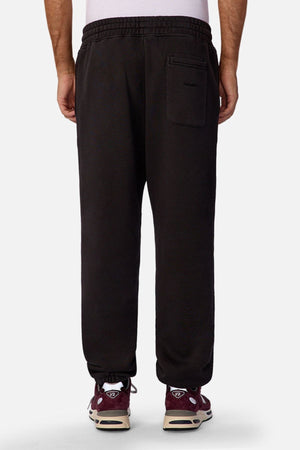 A person faces away, wearing Industrie The New Del Sur Trackpant—relaxed fit black joggers with an elastic waistband and back pocket, paired with maroon and white sneakers. Only their lower half is visible against a plain background—ideal men’s loungewear.