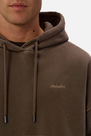 A close-up of a person wearing the Industrie The New Del Sur Washed Hoodie in brown acid wash, featuring drawstrings, "Industrie" chest embroidery, and an oversized fit for a relaxed look. The person’s face is partially visible.