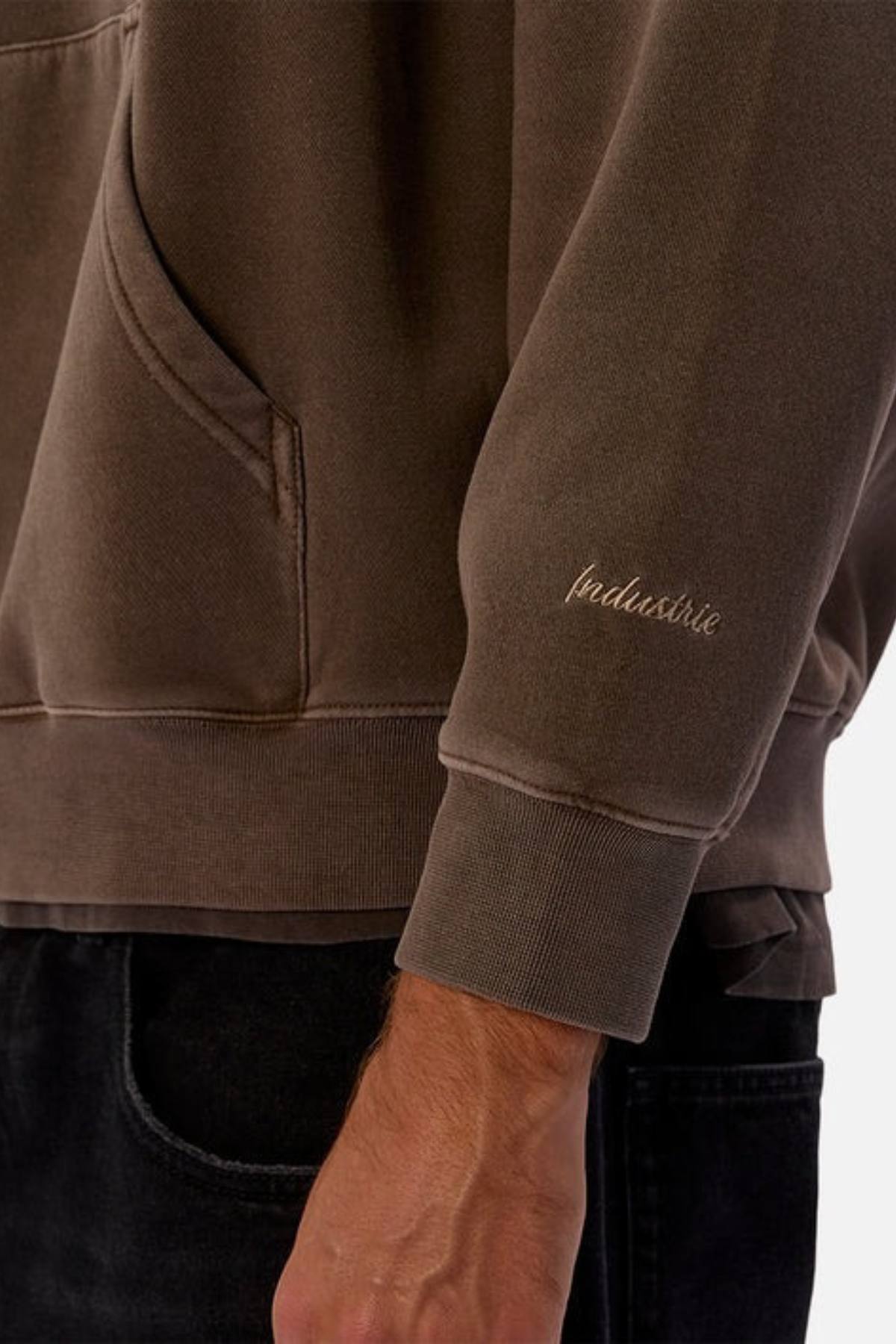 A close-up of a person wearing the Industrie The New Del Sur Washed Hoodie in brown, featuring an oversized fit and front pocket. "Industrie" is embroidered on the left sleeve. It's paired with black pants for a casual style.