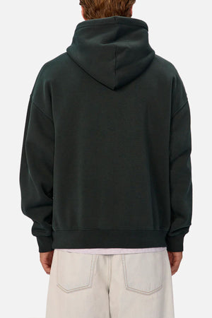 A person is seen from behind wearing the Industrie The New Del Sur Washed Hoodie in dark green with the hood down, paired with light pants. The background is plain and light-colored.