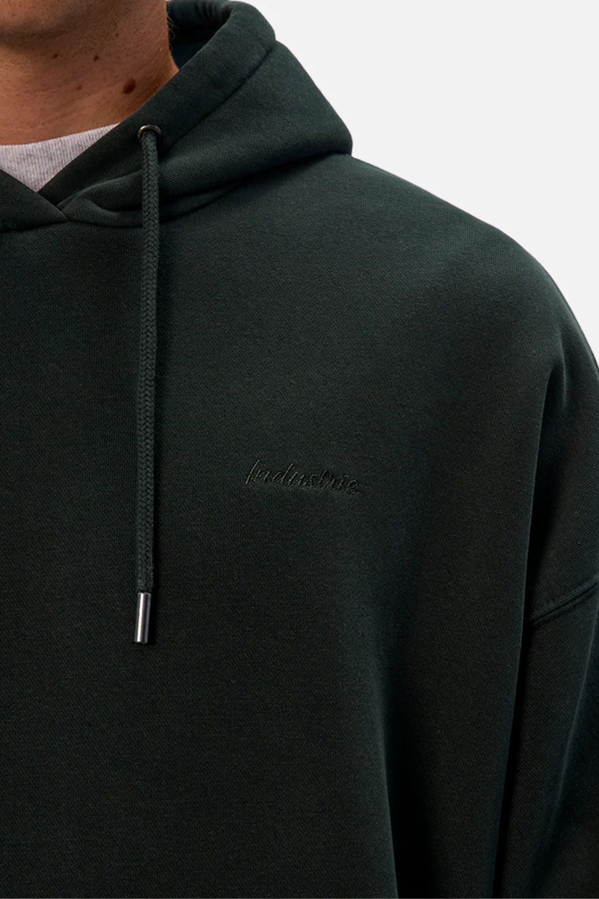 Close-up of a person wearing the INDUSTRIE The New Del Sur Washed Hoodie in dark green, featuring subtle chest embroidery, metal-tipped drawstrings on the hood, and a white shirt visible at the neckline.