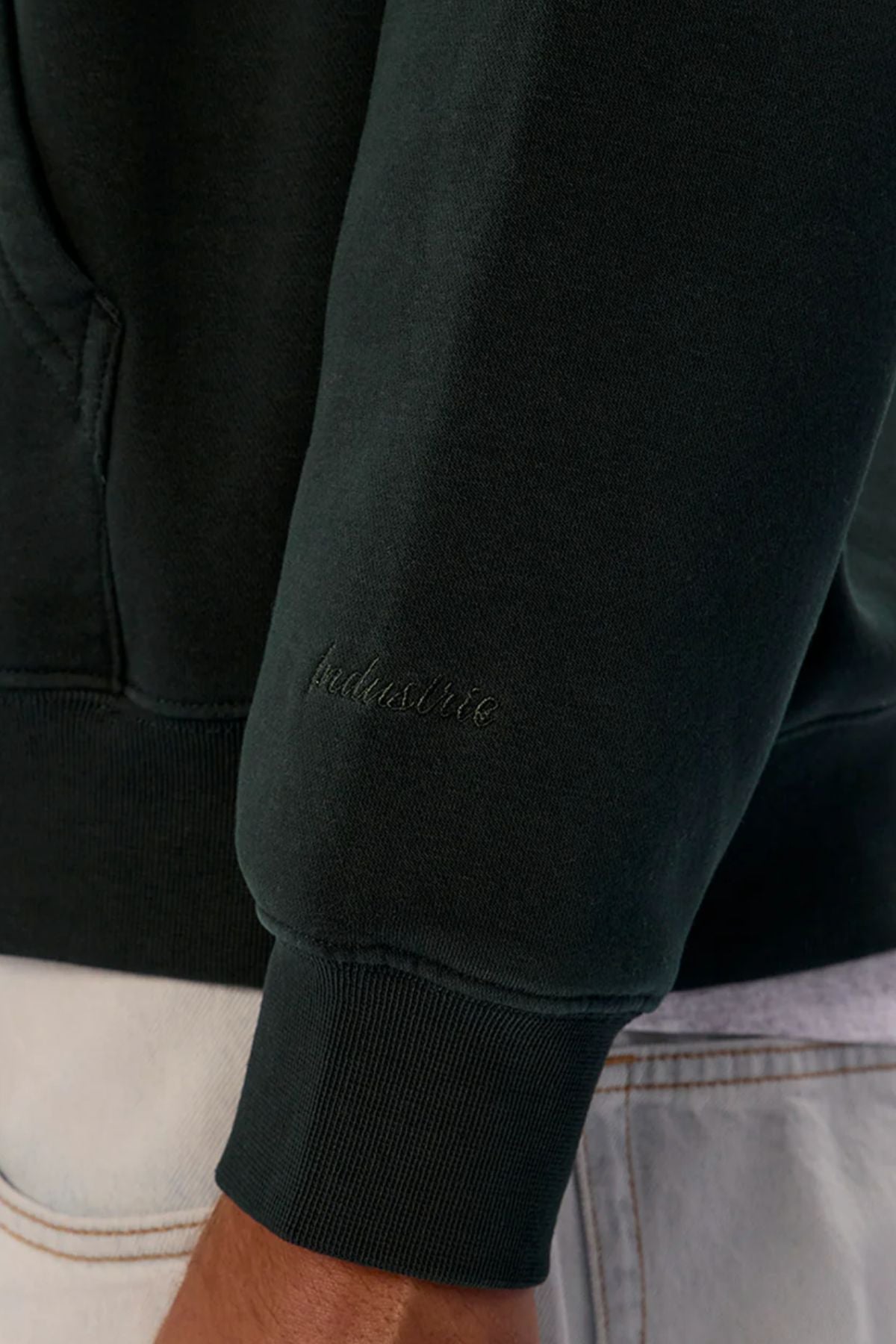 A close-up of the Industrie The New Del Sur Washed Hoodie sleeve in dark green, featuring "inclusive" embroidery in matching thread, shown beside light-colored pants with visible stitching.