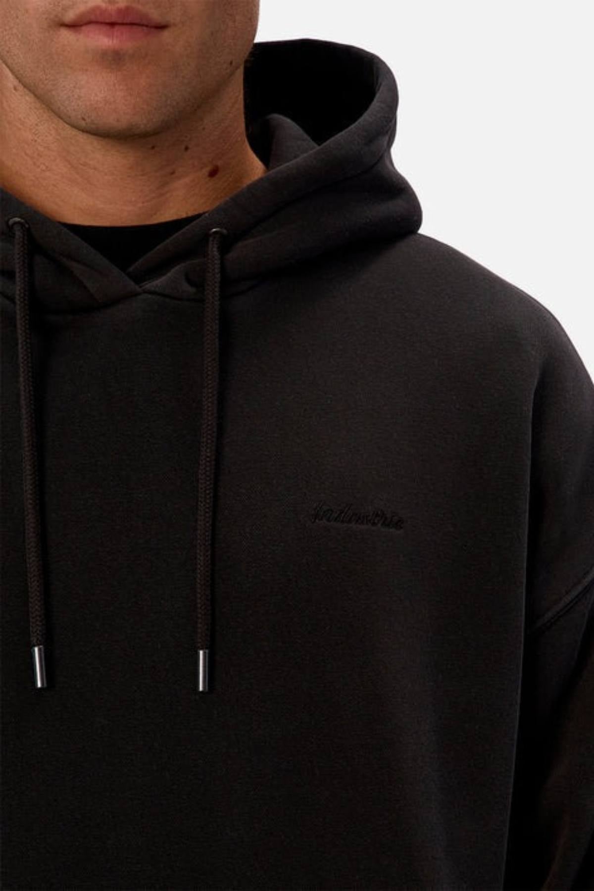 A close-up shows a person wearing the Industrie The New Del Sur Washed Hoodie in black. Only their lower face and upper torso are visible. The oversized hoodie has drawstrings and subtle chest embroidery.