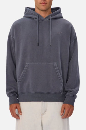 A person wears the Industrie The New Del Sur Washed Hoodie in oversized, faded dark gray with a front pouch pocket and drawstrings, paired with light pants, standing against a plain light background.