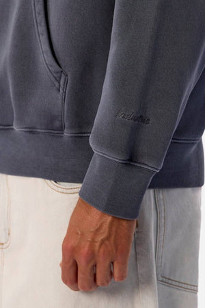 A person wearing the Industrie The New Del Sur Washed Hoodie in an oversized faded dark gray style with sleeve embroidery, paired with light-colored jeans and their hand resting by their side.