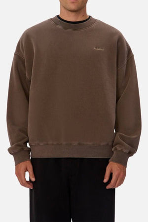 Someone wears an oversized brown Industrie The New Del Sur Washed Sweat with long sleeves and black pants, standing against a plain light background. The sweatshirt has a small embroidered Industrie logo on the left chest.
