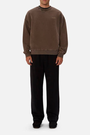 A person stands against a plain background, wearing an oversized Industrie The New Del Sur Washed Sweat and wide-leg black pants with black shoes. Their hands are by their sides and their face is partially visible.