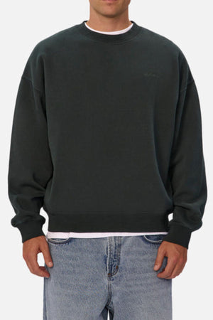 A man wears the Industrie The New Del Sur Washed Sweat in an oversized fit, layered over a white shirt and light blue jeans, standing against a plain, light background. His head is not visible.