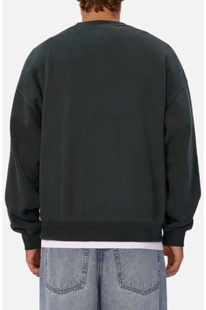 A person wears the Industrie The New Del Sur Washed Sweat in dark green with an oversized fit, styled with light blue jeans and a white shirt peeking from underneath, set against a plain light background.