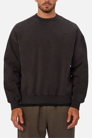 A man wears the Industrie The New Del Sur Washed Sweat, an oversized fit black crew neck sweatshirt with long sleeves and minimal detailing, paired with brown pants, standing against a plain light background.