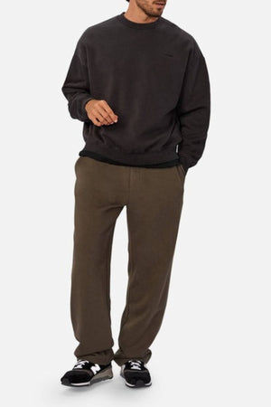 A man stands against a plain white background wearing an oversized Industrie The New Del Sur Washed Sweat, olive green sweatpants, and black New Balance sneakers, with one hand in his pocket.