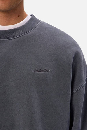 Wearing the Industrie The New Del Sur Washed Sweat in dark grey, styled oversized over a white shirt. The sweatshirt has subtle embroidered chest text. Only the lower face, neck, and upper torso are visible.