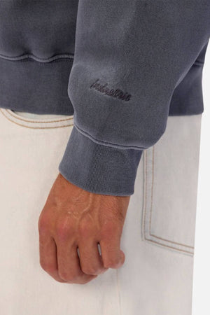 A close-up of a right hand resting on light-colored pants, wearing an oversized fit gray Industrie The New Del Sur Washed Sweat with "Relentless" embroidered near the cuff in small, dark script.