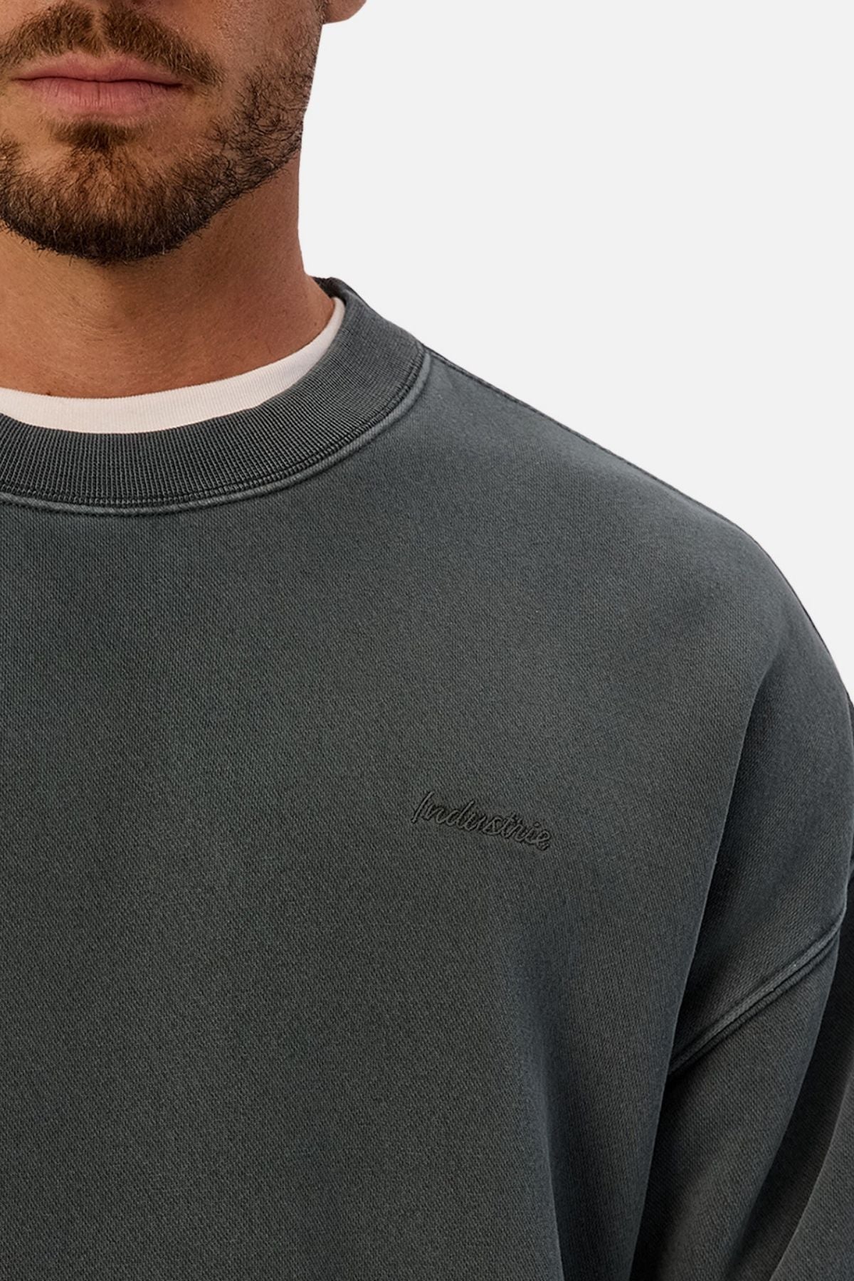 A man wears the Industrie The New Del Sur Washed Sweat in dark gray, featuring subtle chest embroidery and a white tee underneath, shown from the shoulders up against a plain background.
