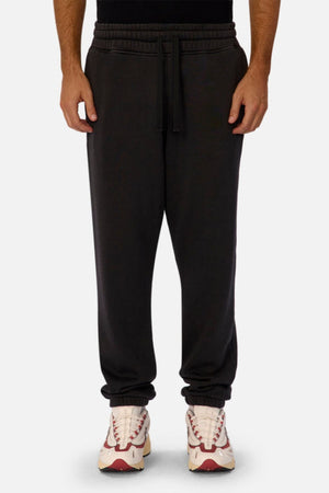 Wearing Industrie The New Del Sur Washed Trackpant with a drawstring waist and elastic cuffs, and white sneakers with red accents, a person stands against a plain light background.