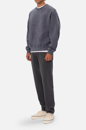 A person stands against a plain light background wearing an oversized dark grey sweatshirt and Industrie The New Del Sur Washed Trackpant with a drawstring waist, layered over a white shirt and paired with white sneakers.
