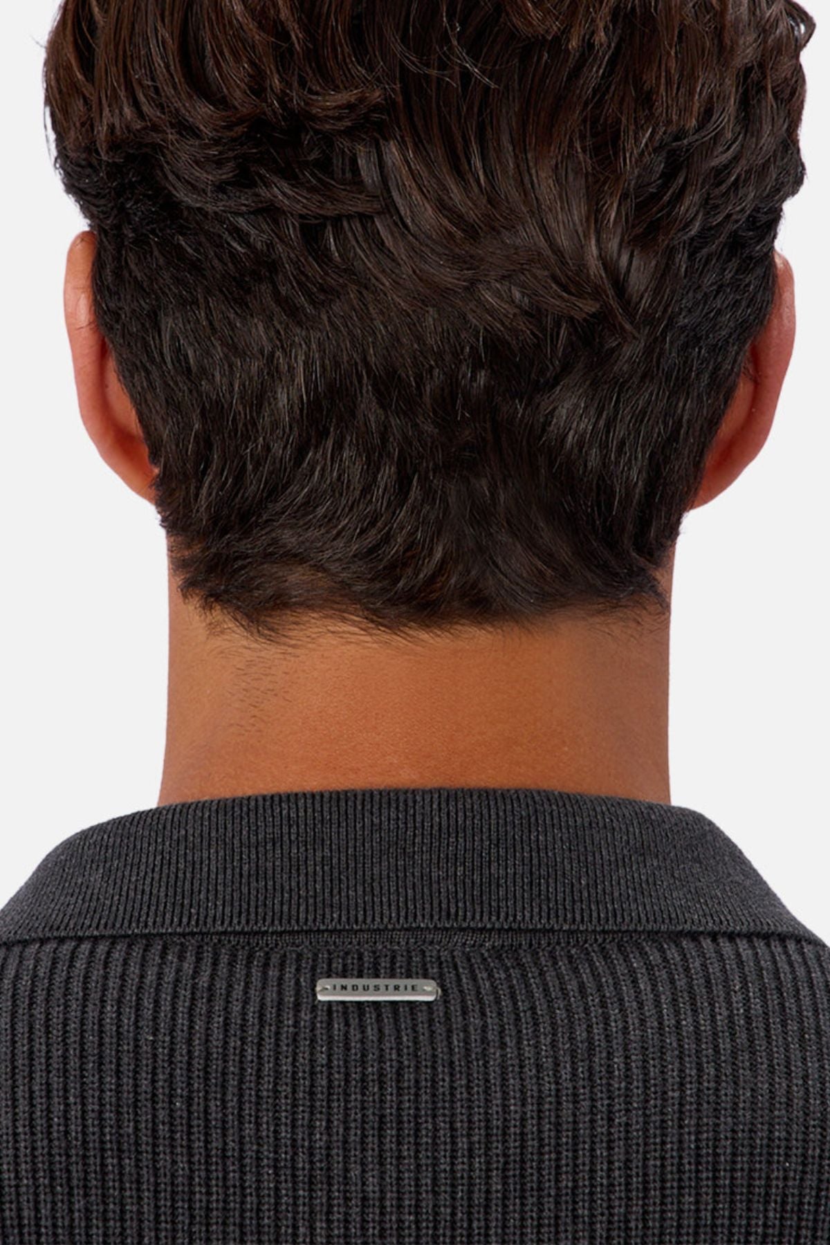 A close-up of the back of a person’s head shows short, wavy brown hair and a dark Industrie The New England Polo Knit sweater with a small metallic label on the collar.