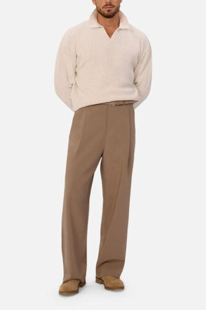 A man stands facing forward in the Industrie The New England Polo Knit, a light beige long-sleeve knit sweater, paired with high-waisted tan belted trousers and light brown shoes. His hands are behind his back against a plain white background.