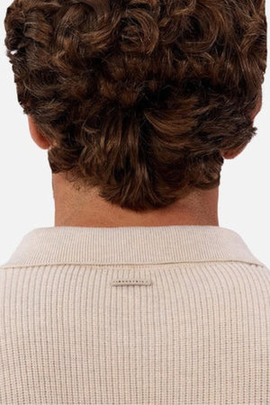 A person with short, curly brown hair is seen from behind wearing the Industrie The New England Polo Knit sweater in cream. The high-quality knit fabric stands out against the plain, light-colored background.