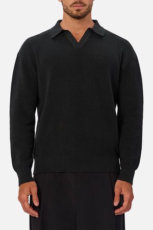 A man wearing the Industrie The New England Polo Knit stands against a plain light gray background. The polo features a V-neck, collar, and high-quality knit fabric. His face is partially visible, and he wears black pants.