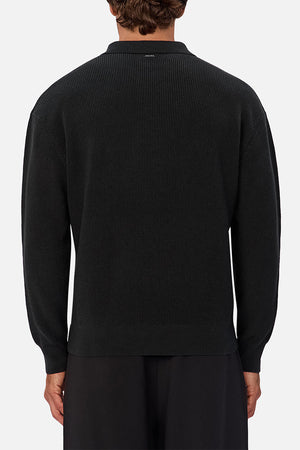A person viewed from behind wears the Industrie The New England Polo Knit, a black long-sleeve knit sweater, styled with loose black pants against a simple light gray background.