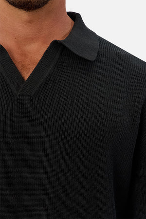Close-up of a person wearing the Industrie The New England Polo Knit, a dark knit sweater featuring a collar and V-neck, shown from shoulders to upper chest against a plain light background.