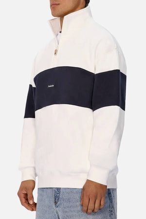 Wearing the Industrie The New Luton Track Top—a white men's top with a navy stripe, chest logo, and stand-up collar—paired stylishly with light blue jeans.