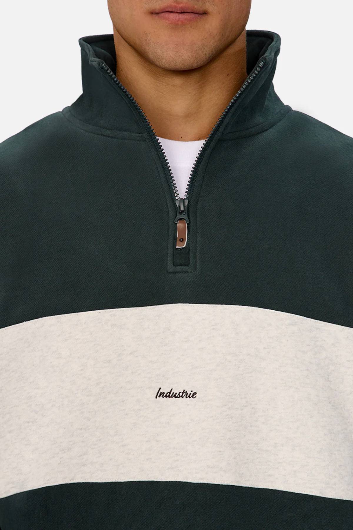 A person wears the Industrie The New Luton Track Top, a dark green quarter zip with a relaxed fit, white chest stripe, and "Industrie" embroidered at the center. Only the lower face is shown.