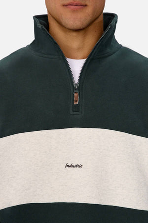 A person wears the Industrie The New Luton Track Top, a dark green quarter zip with a relaxed fit, white chest stripe, and "Industrie" embroidered at the center. Only the lower face is shown.