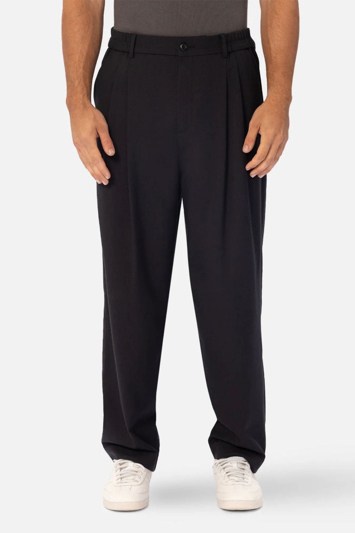 Showcasing a professional look, a person wears Industrie The New Milano Pant—loose-fit black pleated trousers—paired with a tucked-in dark shirt and white sneakers, against a plain light background. The image is cropped from shoulders to feet.