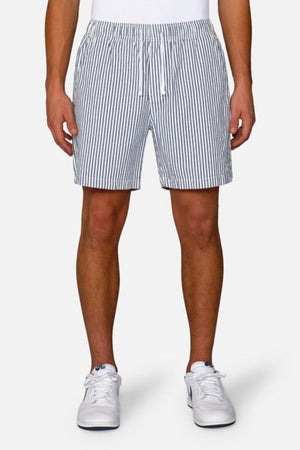 A person stands against a plain white background, sporting the Industrie The New Southwest Short, featuring blue and white vertical stripes and an elasticated waistband. They complete the look with crisp white socks and sneakers.