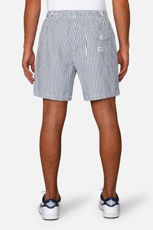 Rear view of a person wearing blue and white Industrie The New Southwest Short, featuring an elasticated waistband, paired with white socks and sneakers with blue accents, standing against a plain background.
