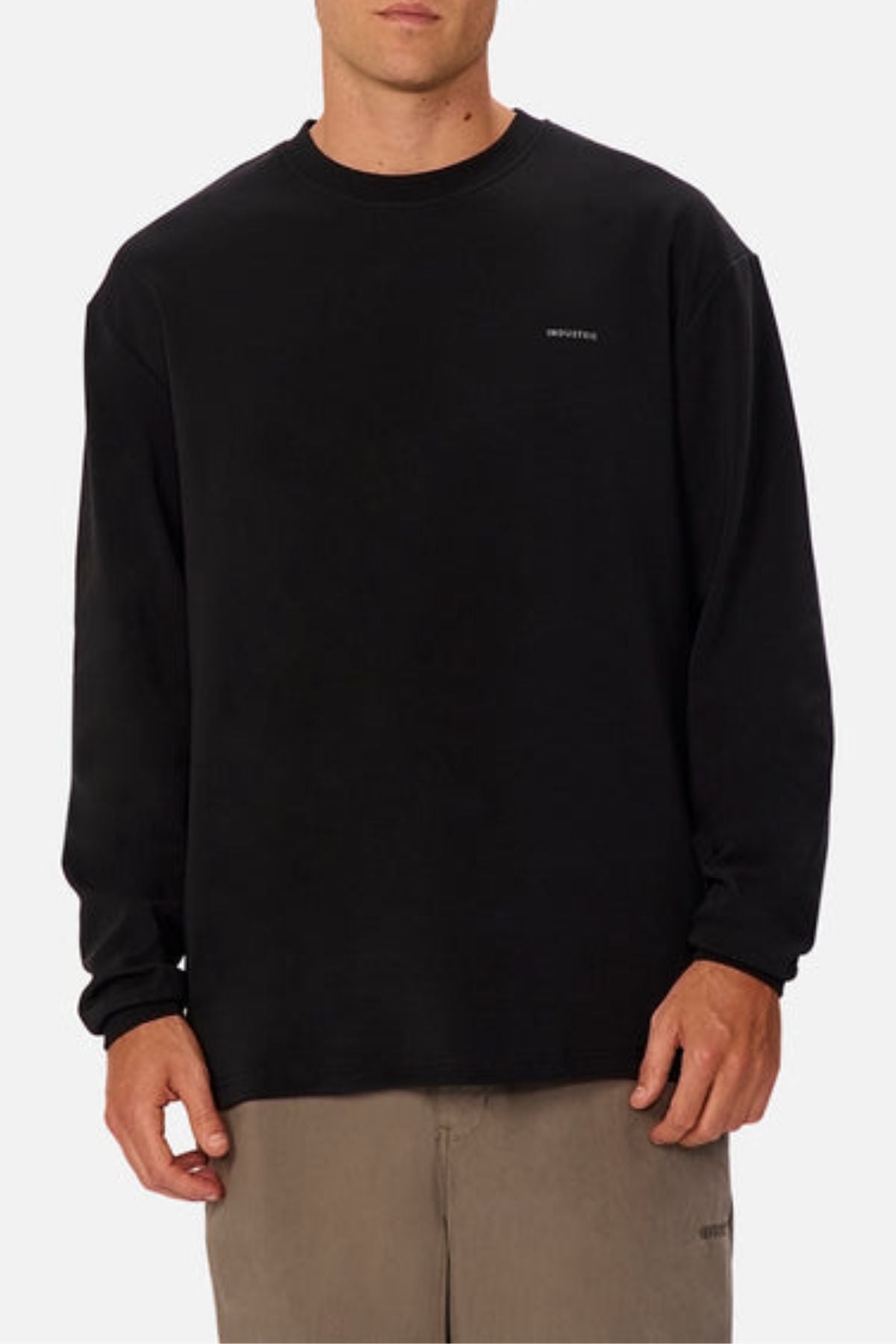 A person stands against a white background wearing an oversized fit black Industrie The Newport Long Sleeve Tee with small white chest text and light brown pants.