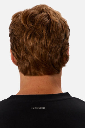 The image shows the back view of a person with short, wavy brown hair wearing an oversized black Industrie The Newport Long Sleeve Tee, featuring "INDUSTRIE" printed in white on the upper back.