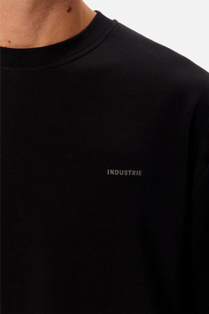 Close-up of a person wearing the Industrie The Newport Long Sleeve Tee, an oversized black crewneck with "INDUSTRIE" printed in small gray letters on the left chest. Only the lower neck and upper chest are visible.