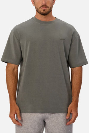 A man wears the Industrie The Newport Tee, showcasing an oversized fit and heavyweight fabric. He stands facing forward in plain gray and light gray shorts against a white background with his arms relaxed at his sides.