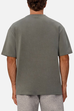 A man is shown from the back wearing Industrie The Newport Tee in a grayish-green, oversized fit with light gray shorts against a plain, light background.