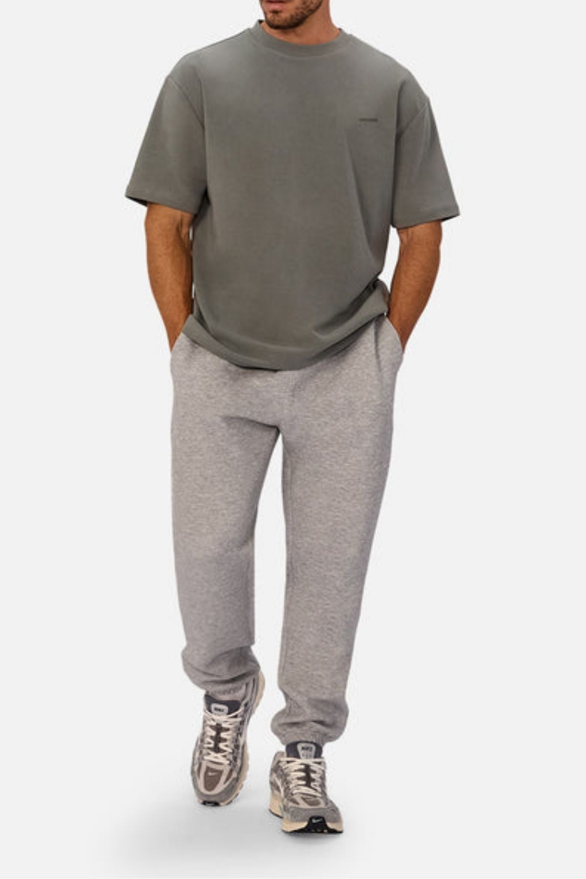 A man wears the Industrie The Newport Tee in a loose, oversized fit with light gray sweatpants and gray and white sneakers, standing with his hands in his pockets against a plain white background.