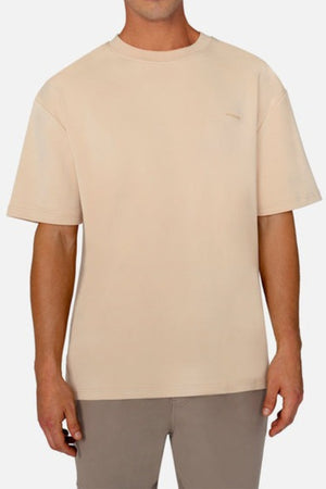 A person wearing an oversized, 100% cotton beige T-shirt stands against a neutral background. The loose-fitting, short-sleeved tee is the Industrie The Newport Tee and has an Industrie branded logo, while the person's arms are relaxed at their sides.