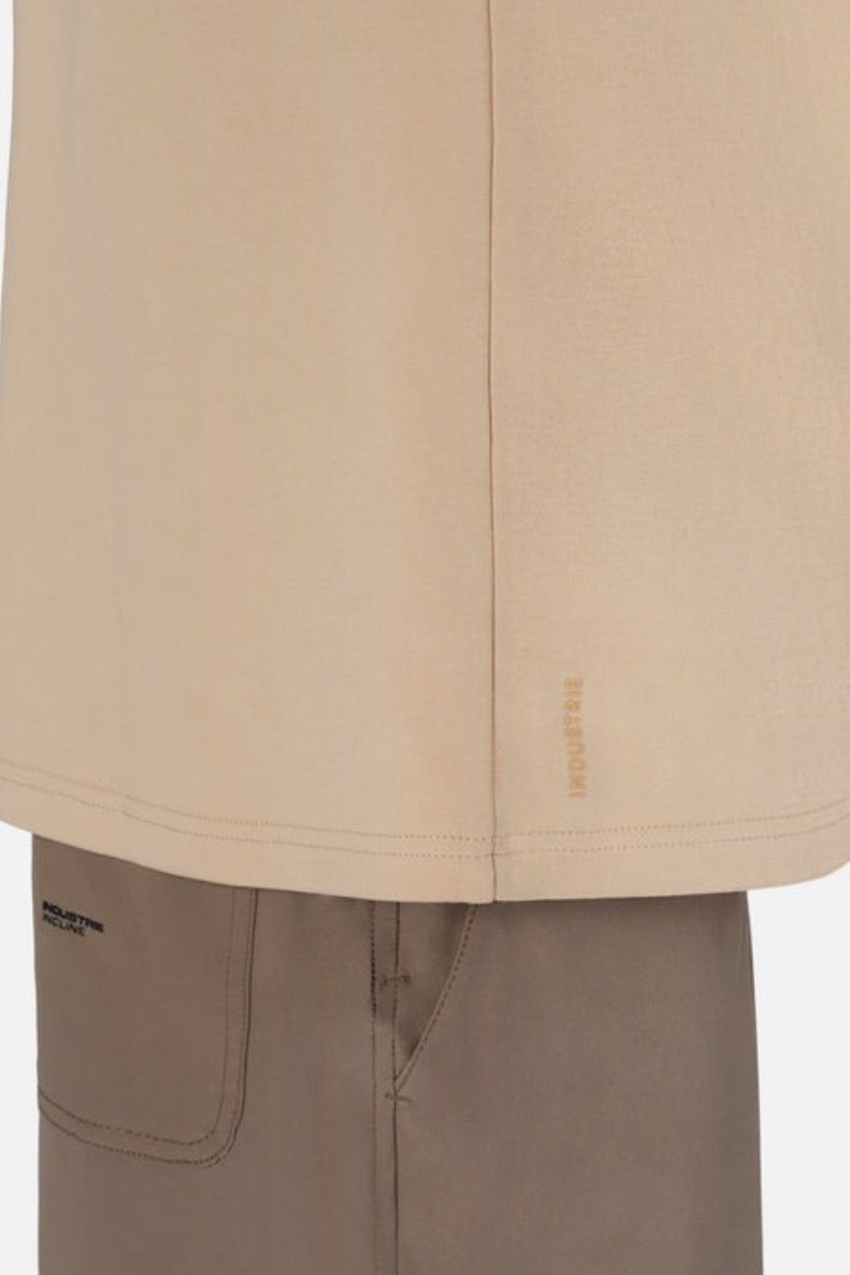 Close-up of a person wearing the oversized fit Industrie The Newport Tee in beige, crafted from 100% cotton, with "REPRESENT" on the side hem. It is paired with brown pants featuring visible stitching, a pocket detail on the back, and Industrie’s distinctive branded logo.