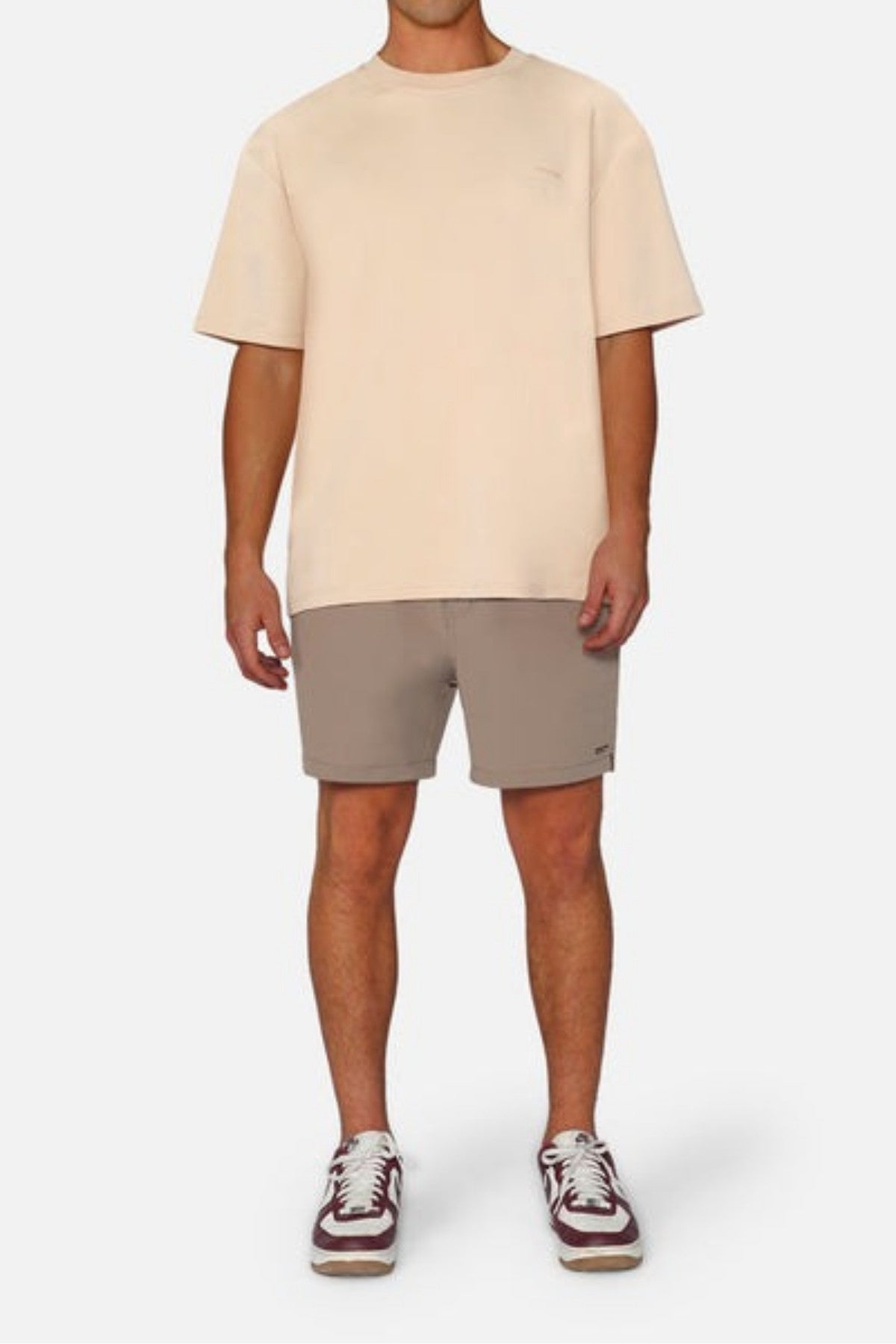 A person in an oversized fit beige Industrie The Newport Tee, gray shorts, and maroon and white sneakers stands against a plain white background.