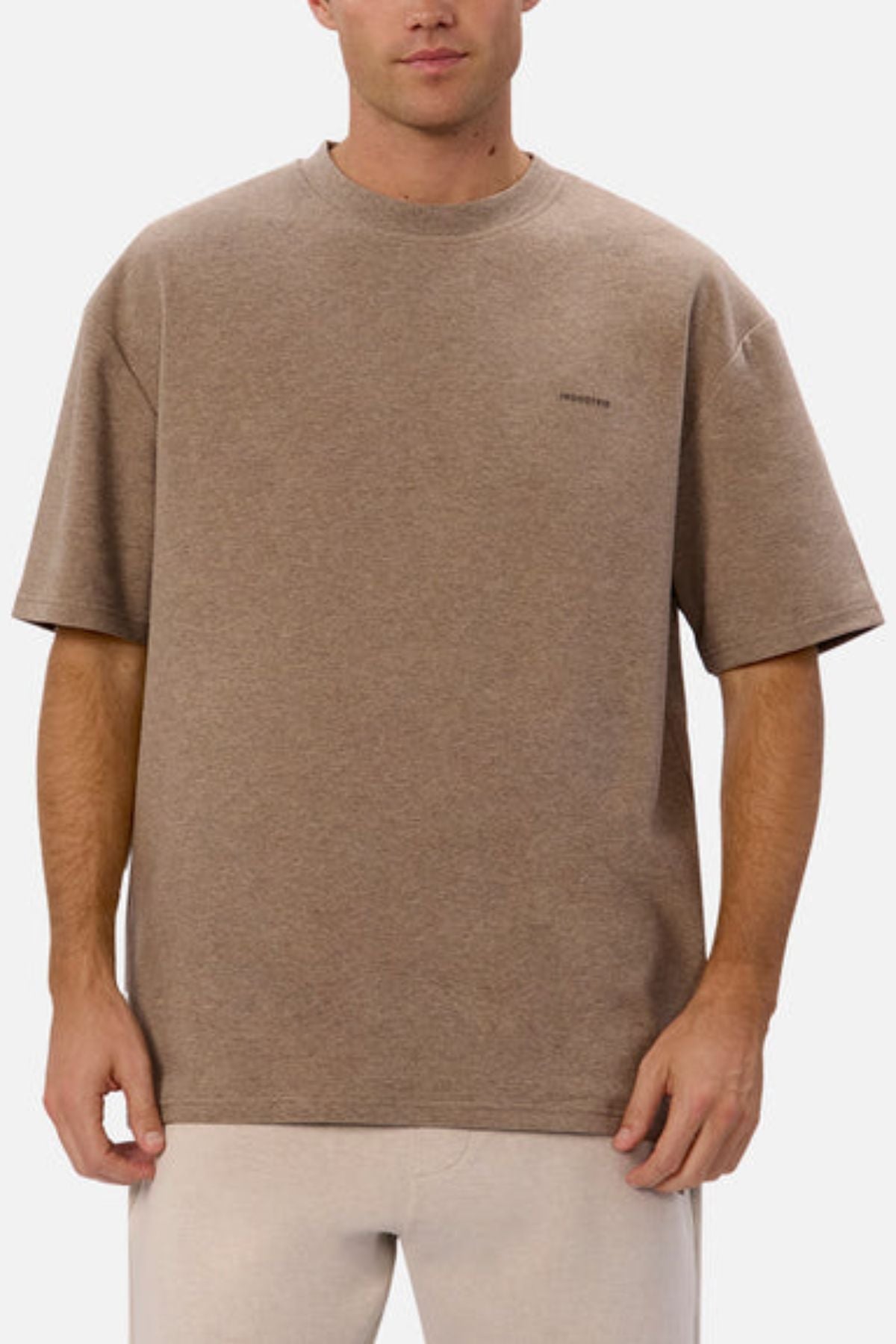 A person is wearing an oversized fit, brown T-shirt and light-colored pants. The T-shirt, which is likely Industrie The Newport Tee, has a small logo on the chest and is crafted from 100% cotton. The background is plain white.