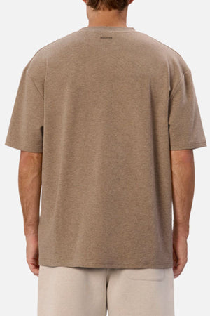A person wearing Industrie's The Newport Tee in brown paired with light-colored shorts is turned away from the camera. The oversized fit of this 100% cotton tee enhances a relaxed vibe against the simplistic white backdrop.