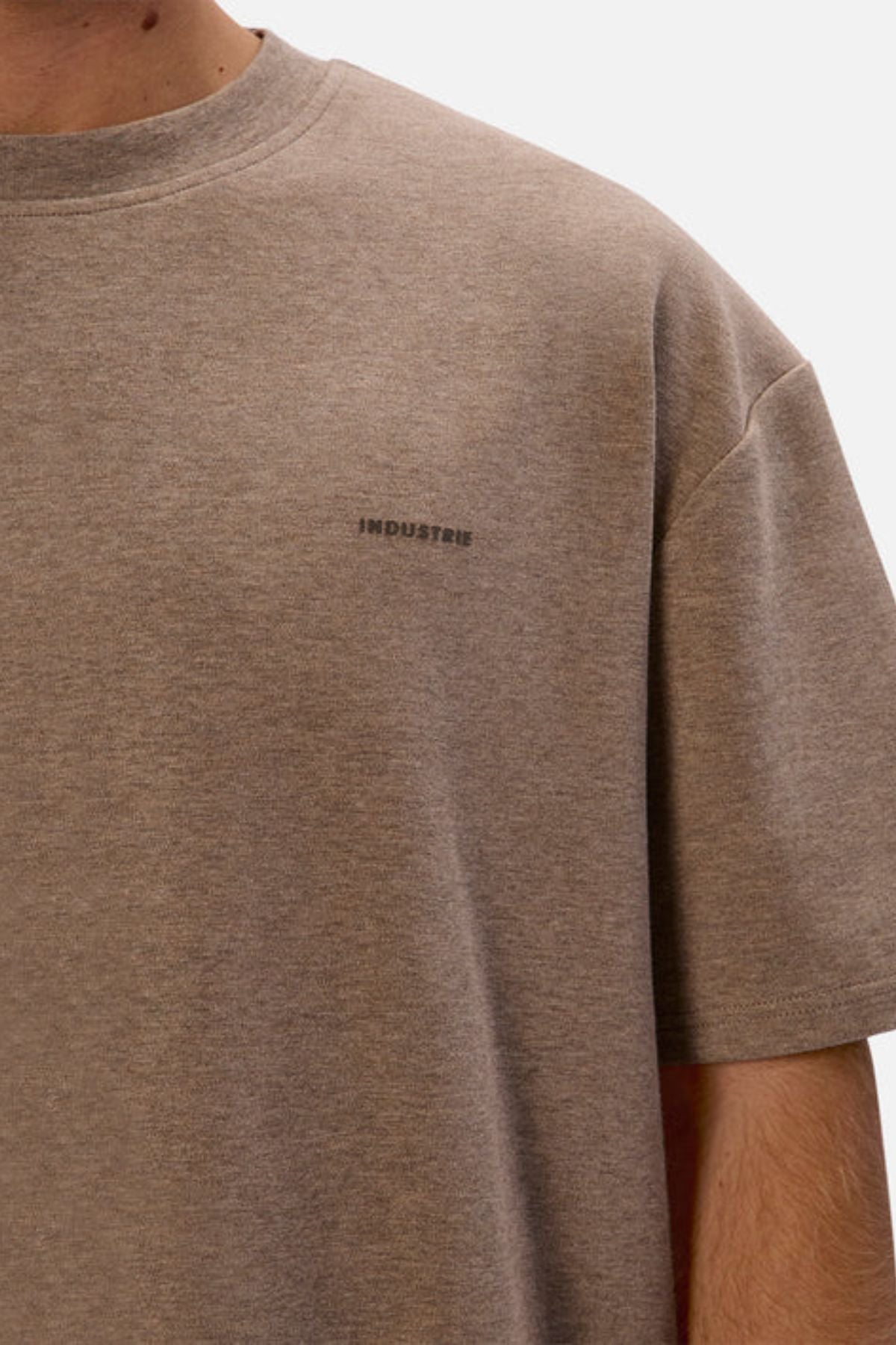 A person wearing Industrie The Newport Tee, featuring a light brown hue and short sleeves, with "INDUSTRIES" subtly printed in small dark letters on the chest. Crafted from 100% cotton, its oversized fit provides a relaxed appearance against a plain white background.