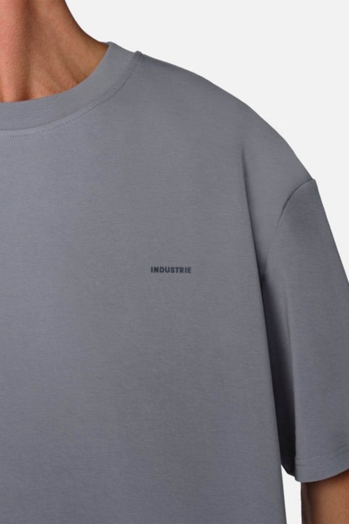 A person wearing an oversized fit Industrie The Newport Tee, made from 100% cotton, showcases the word "INDUSTRIE" printed in small letters on the chest against an off-white background.